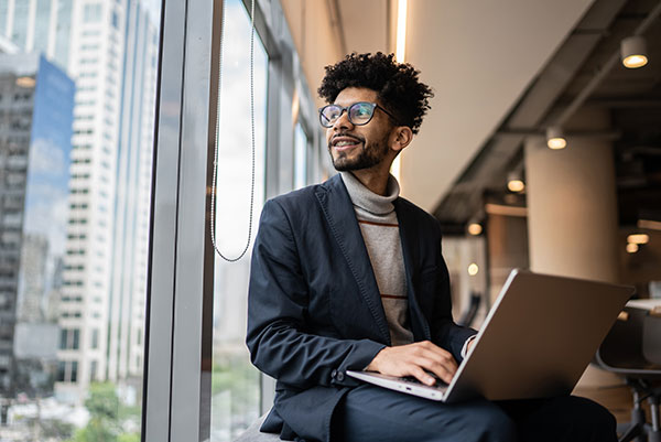 A man with glasses glances cheerfully out a window while typing on a laptop