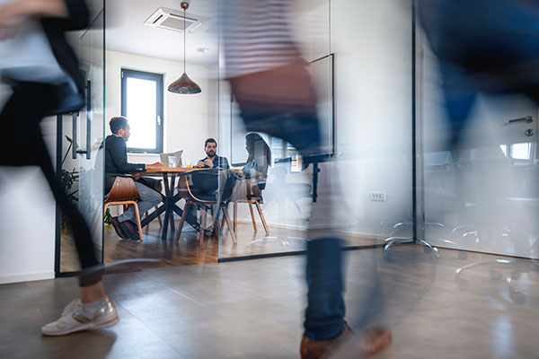 Members of a creative team meet in a glass-walled conference room in a busy office